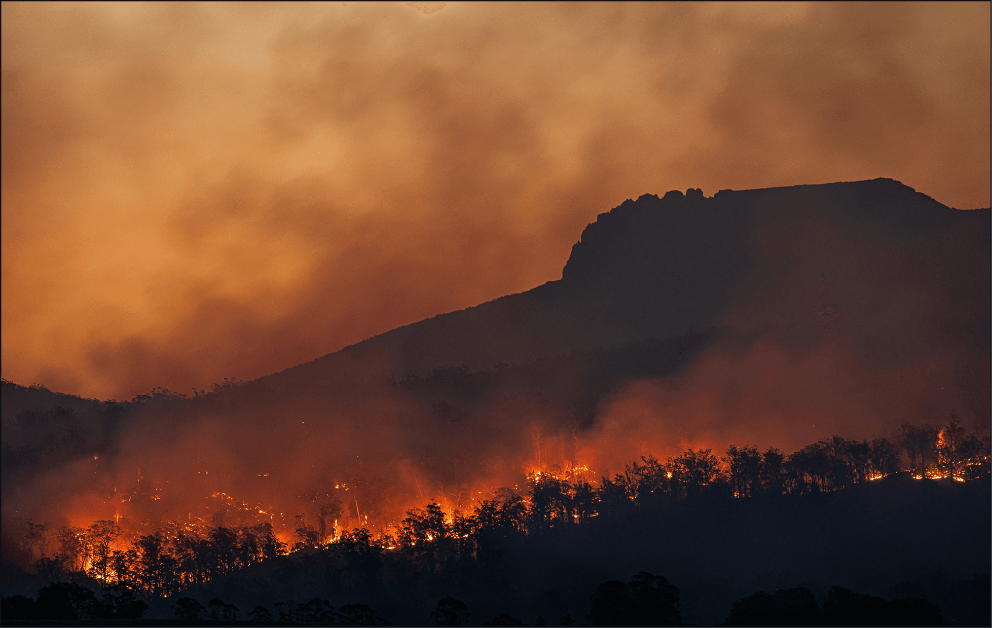 An Australian bushfire at dusk: a dark mountain ridge under heavy orange smoke with a line of burning trees glowing along the slope.