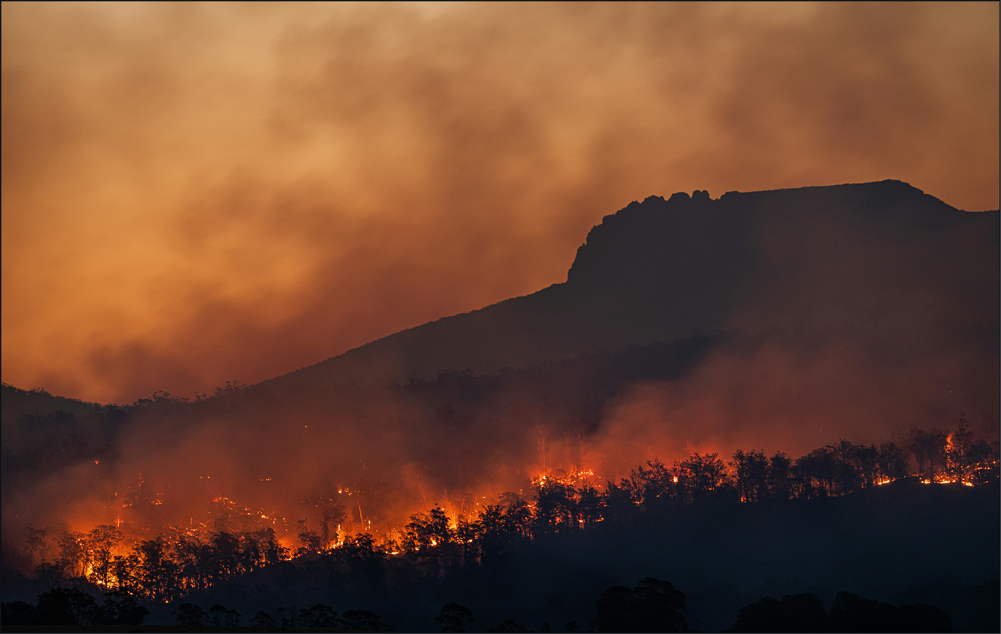An Australian bushfire at dusk: a dark mountain ridge under heavy orange smoke with a line of burning trees glowing along the slope.